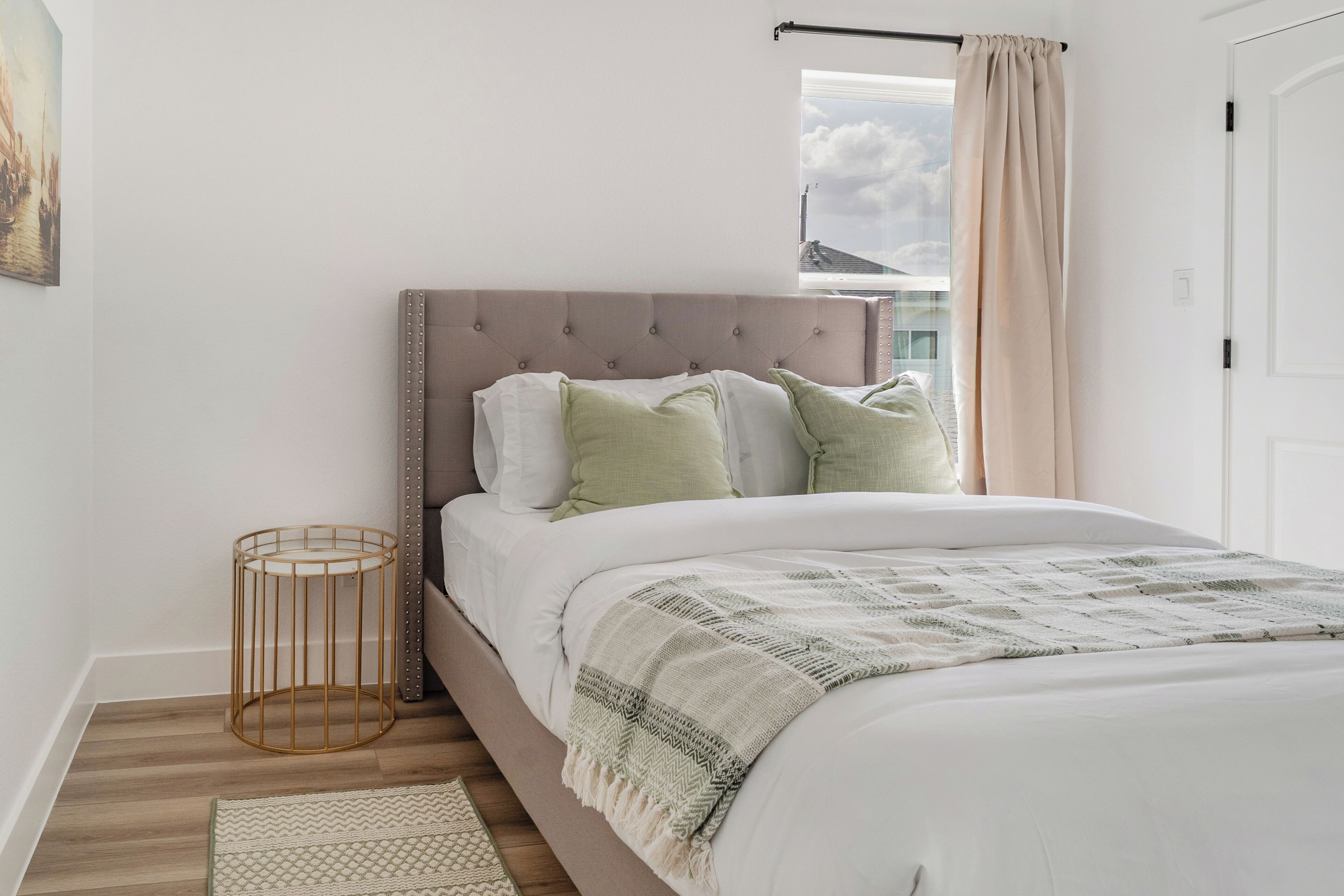 A bedroom with a bed, pillows, a bedside table, a window with curtains, and a wall-mounted picture.