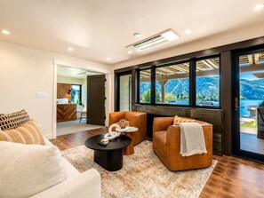Ein modernes Wohnzimmer mit einer Couch, Sesseln, einem Couchtisch und einem Kamin. Durch große Fenster ist ein Blick auf einen See und Berge möglich.