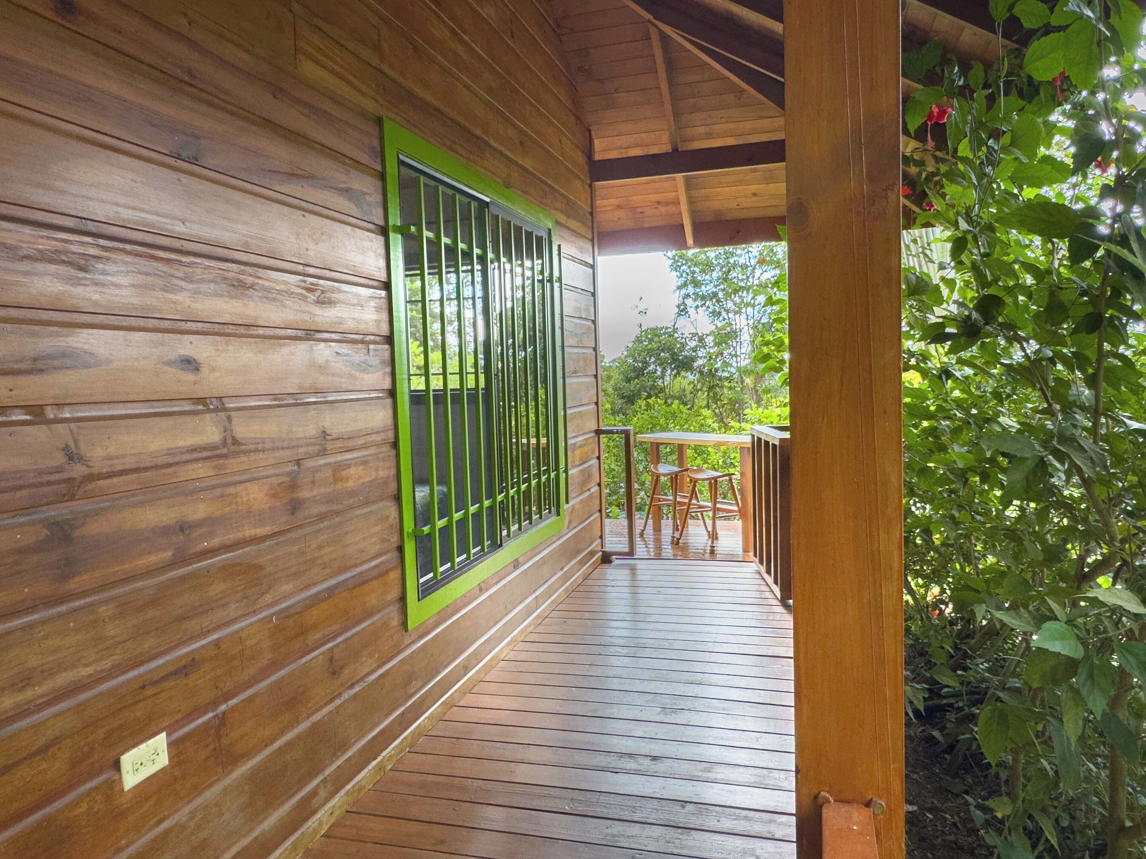 Une terrasse en bois avec une rambarde, une fenêtre avec des barreaux verts et une vue sur la verdure.