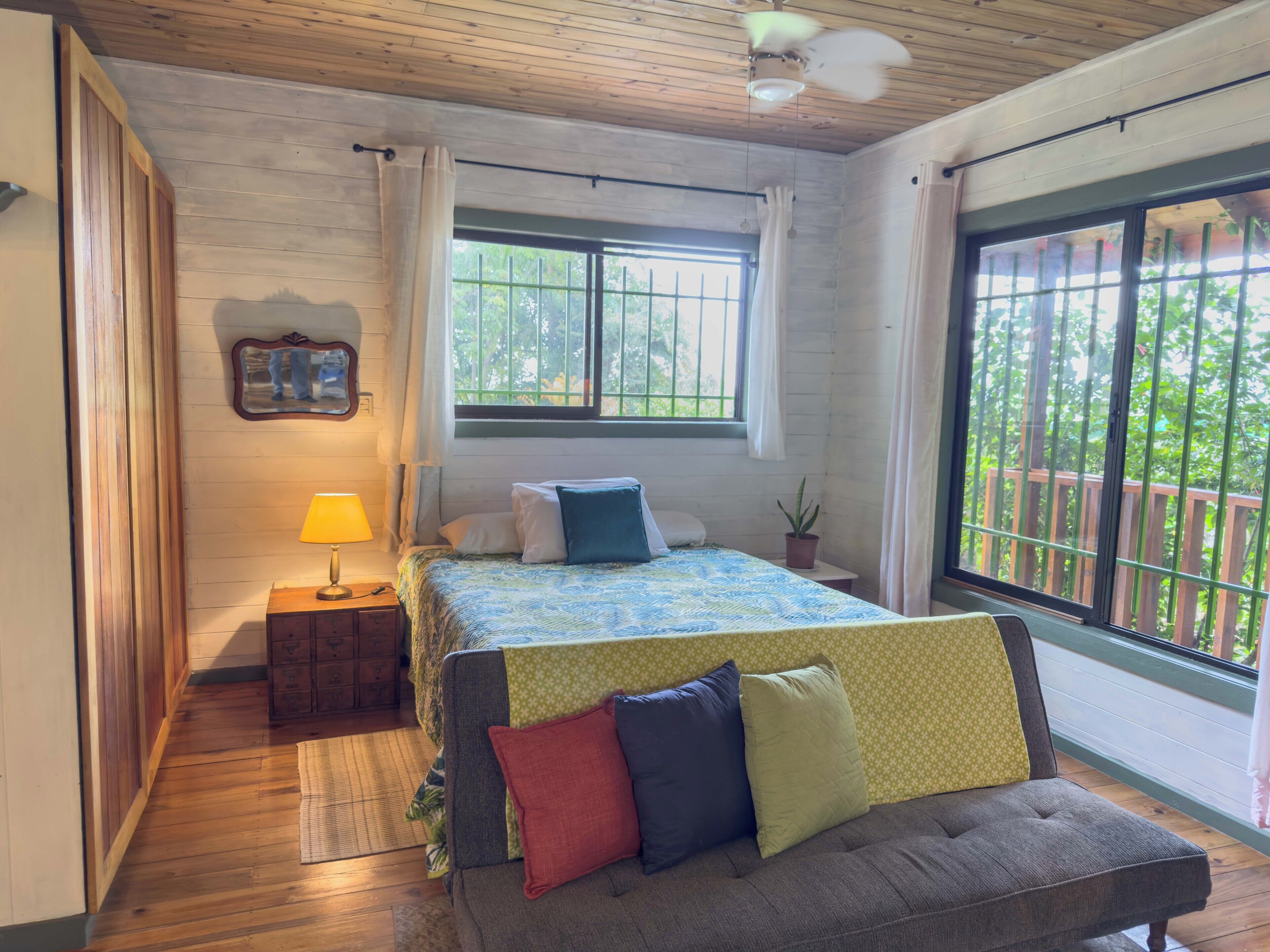 A bedroom with a bed, a bedside table with a lamp, a window with curtains, and a view of greenery outside.