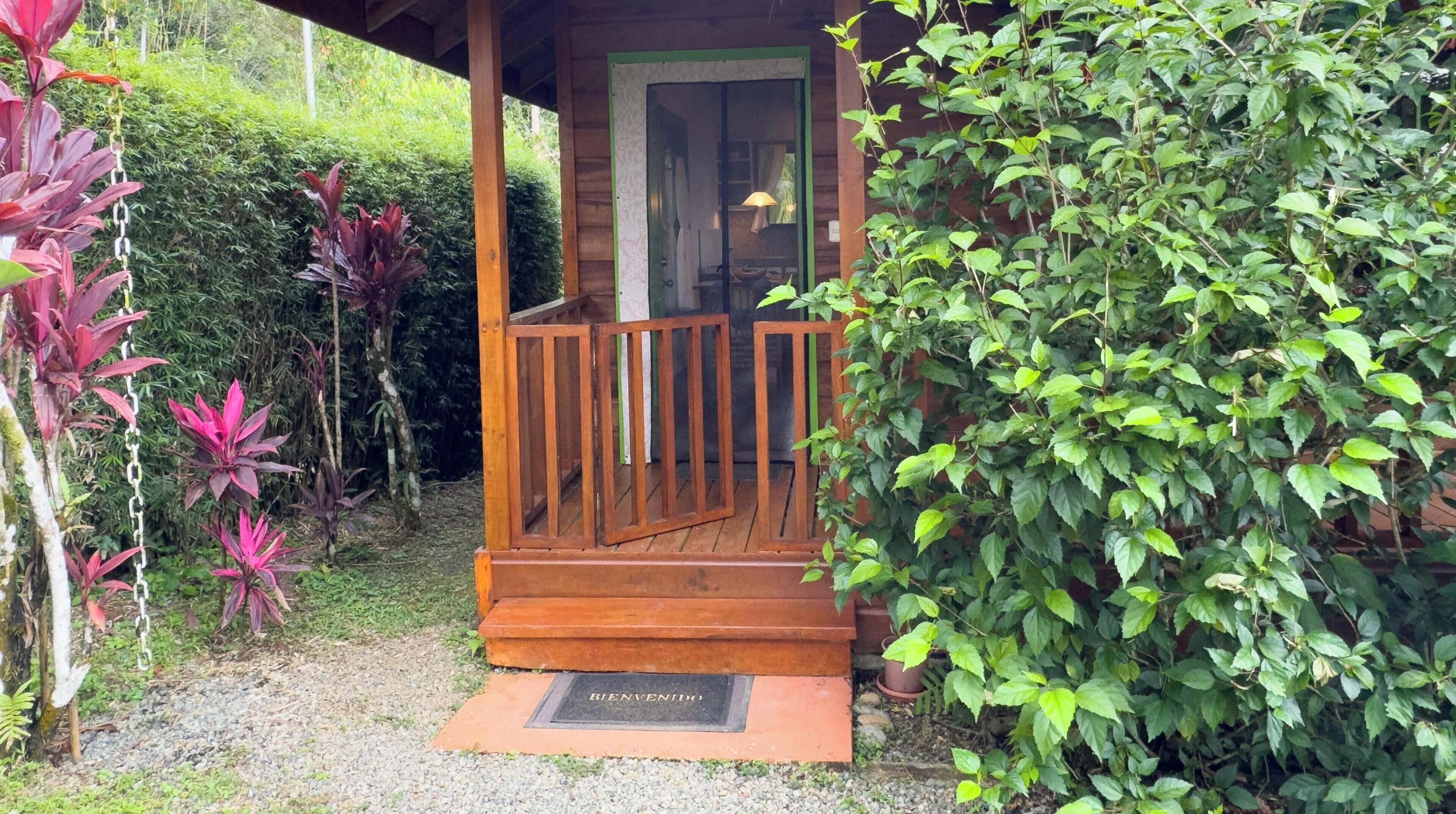 A wooden porch with a glass door, surrounded by lush greenery and a gravel path leading to a welcome mat.