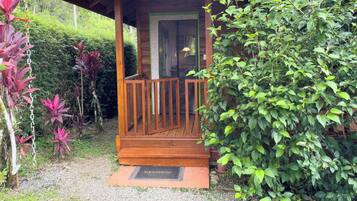 A wooden porch with a glass door, surrounded by lush greenery and a gravel path leading to a welcome mat.
