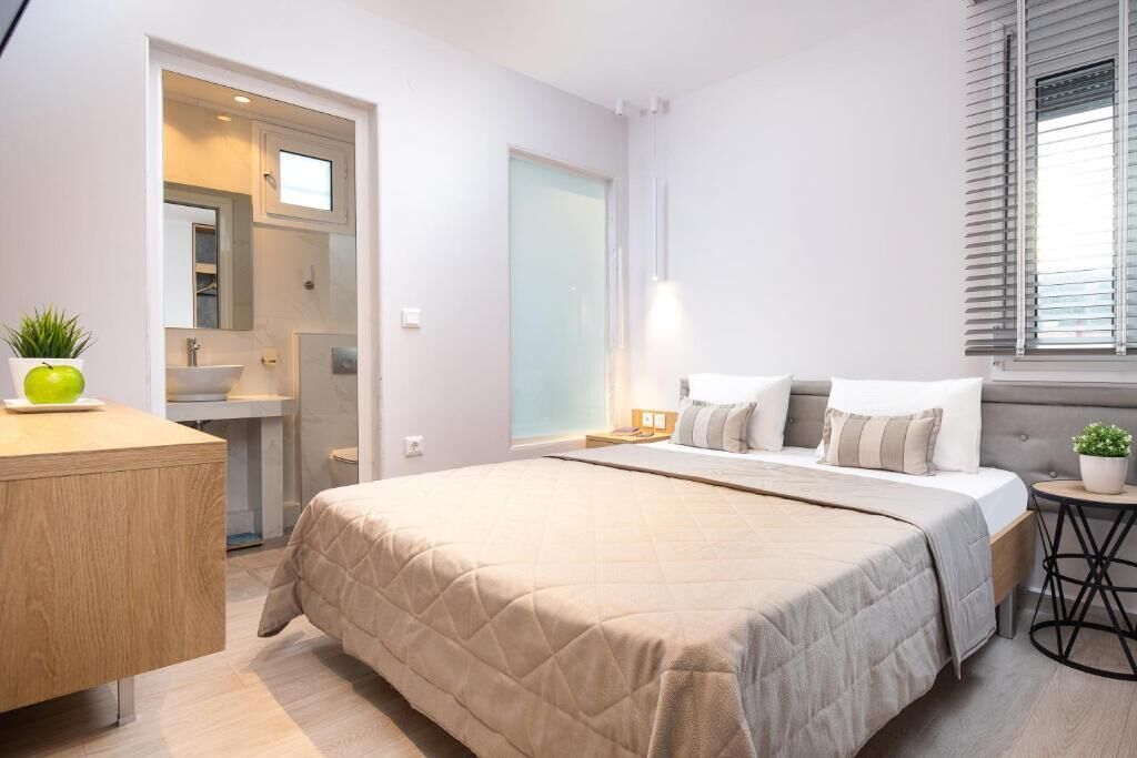 A modern bedroom with a large bed, bedside tables, a small round table, a mirror, and a window with blinds.