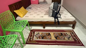 A bedroom with a bed, a green chair, a small table, and a door. - Smriti Homestay (Varanasi)