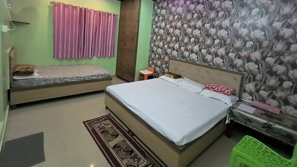 A bedroom with a bed, a nightstand, a small table, a green stool, a ceiling fan, and a wall with a floral wallpaper. - Smriti Homestay (Varanasi)