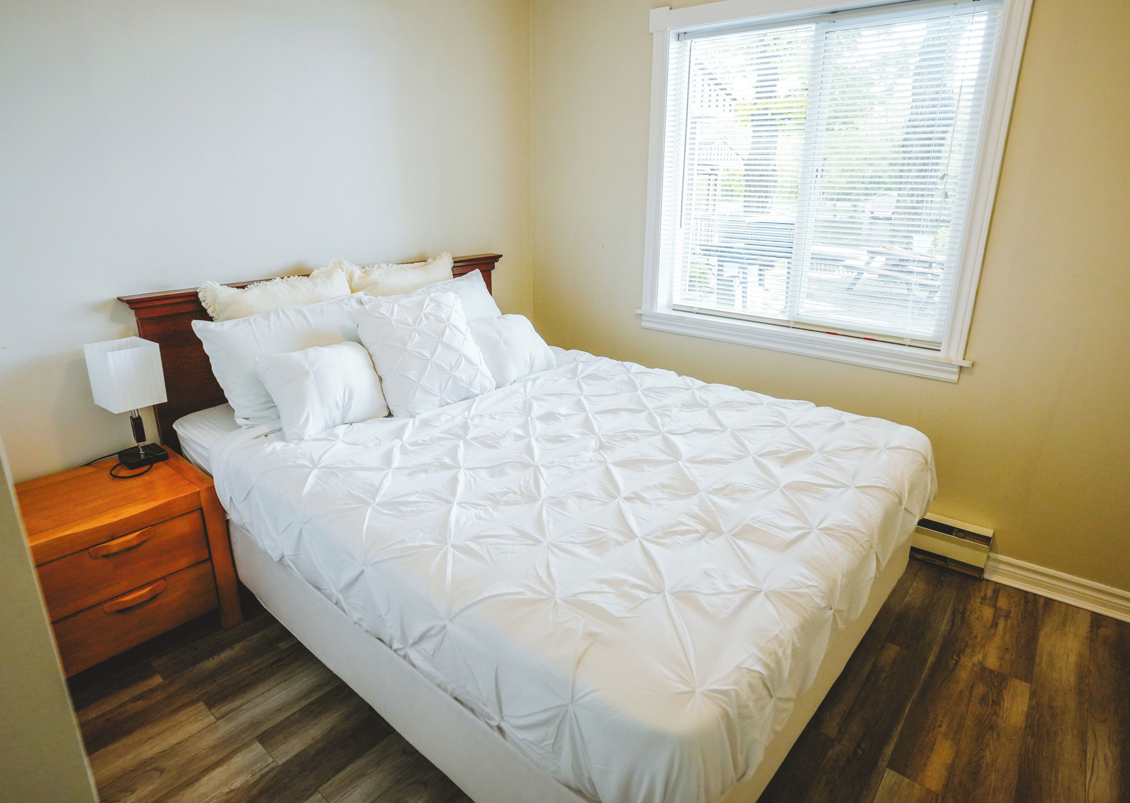 Una cama bien hecha con cabecera de madera, una mesita de noche con lámpara y una ventana con persianas.