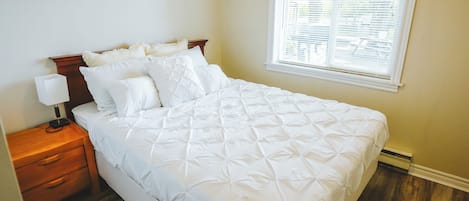 Una cama bien hecha con cabecera de madera, una mesita de noche con lámpara y una ventana con persianas.