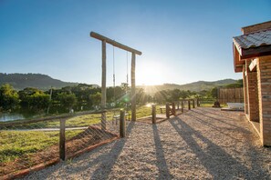 A gravel patio with a swing set and a wooden structure, overlooking a body of water and hills.