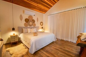 A bedroom with a wooden ceiling, a bed with white bedding, a nightstand, a wall-mounted air conditioner, and a large window with white curtains. - Chalés Ipês da Serra (Serra Negra)