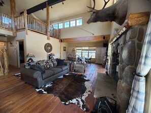A spacious living room with a high ceiling, a stone fireplace, a mounted deer head, and a large window providing natural light. - NEW LISTING!  Peaceful 4-bedroom home in Big Sky ready for your next adventure. (Big Sky)