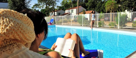 Zona junto a la piscina con una persona leyendo un libro, piscina y una valla blanca.