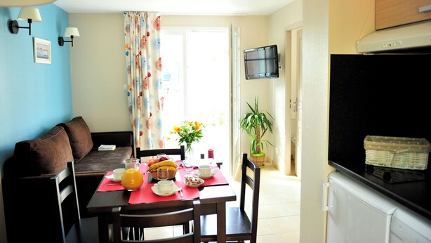 A dining area with a table set for two, a sofa, a TV mounted on the wall, and a potted plant.