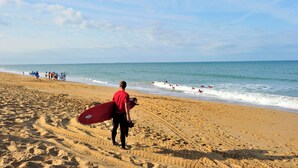 A sandy beach with footprints leading to the water, a person carrying a red surfboard, and a group of people in the distance.