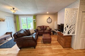 A living room with a brown leather sofa, a wooden cabinet with a TV, and a decorative wall piece. - Rez de Jardin at Magog: Waterfront•Pool•Hot tub•3 rooms (Magog)