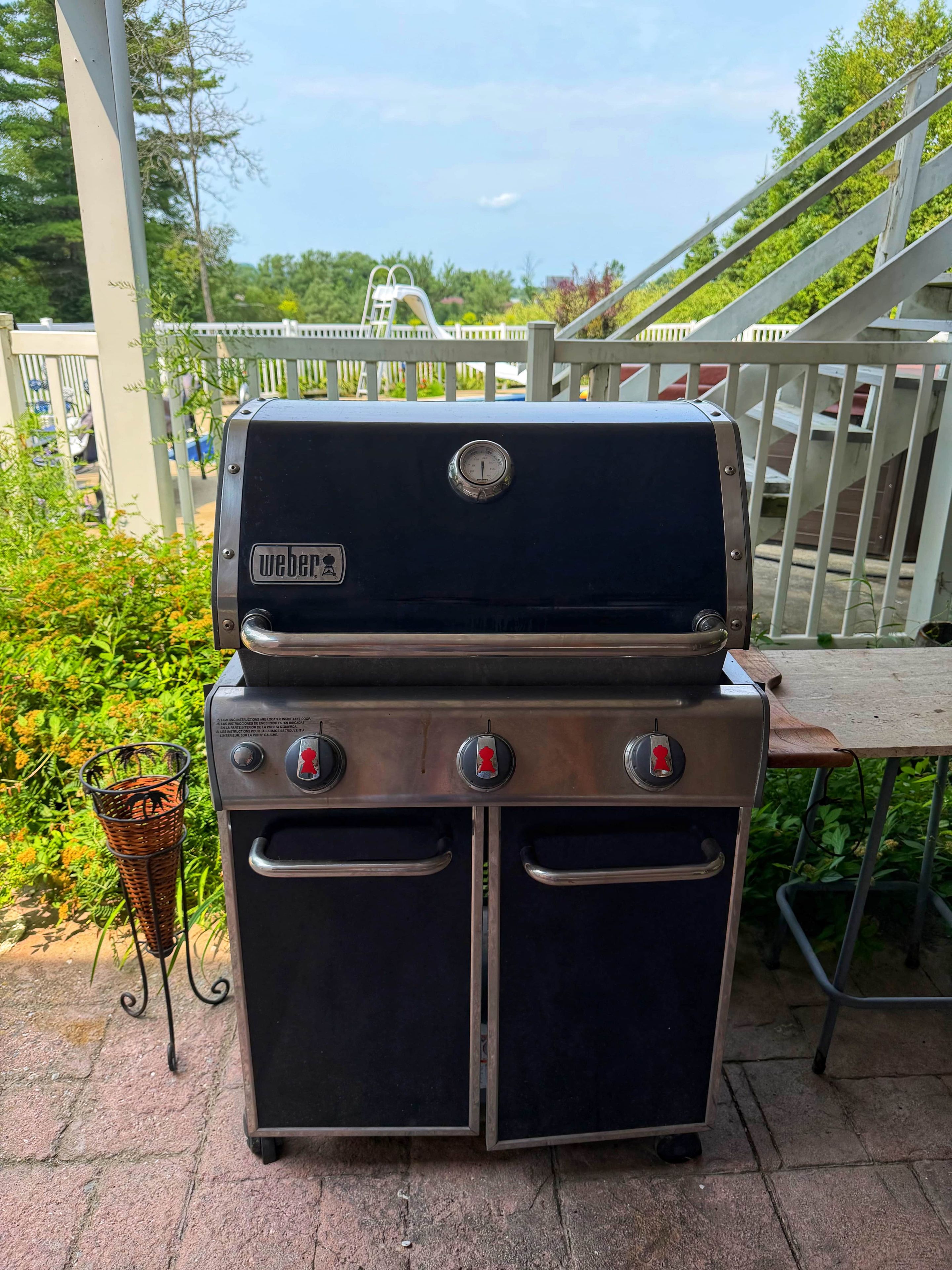 Una parrilla de gas Weber con dos quemadores laterales y una tapa, ubicada al aire libre en un patio con barandilla y follaje de fondo.