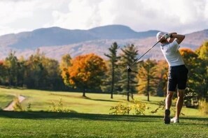 Eine Person spielt Golf auf einem Platz, im Hintergrund Bäume und Berge.