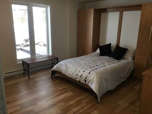 A bedroom with a bed, wooden wardrobe, bench, and a view through a sliding door. - Rez de Jardin at Magog: Waterfront•Pool•Hot tub•3 rooms (Magog)