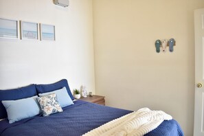 A bedroom with a bed, pillows, a nightstand, a wall-mounted shoe rack, and a door. - Coastal Home San Miguel (San Miguel)