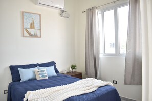 A bedroom with a bed, pillows, a bedside table, a plant, a window with curtains, and a painting on the wall. - Coastal Home San Miguel (San Miguel)