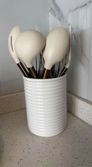 A white ceramic container with handles, filled with metal utensils, placed on a speckled countertop.