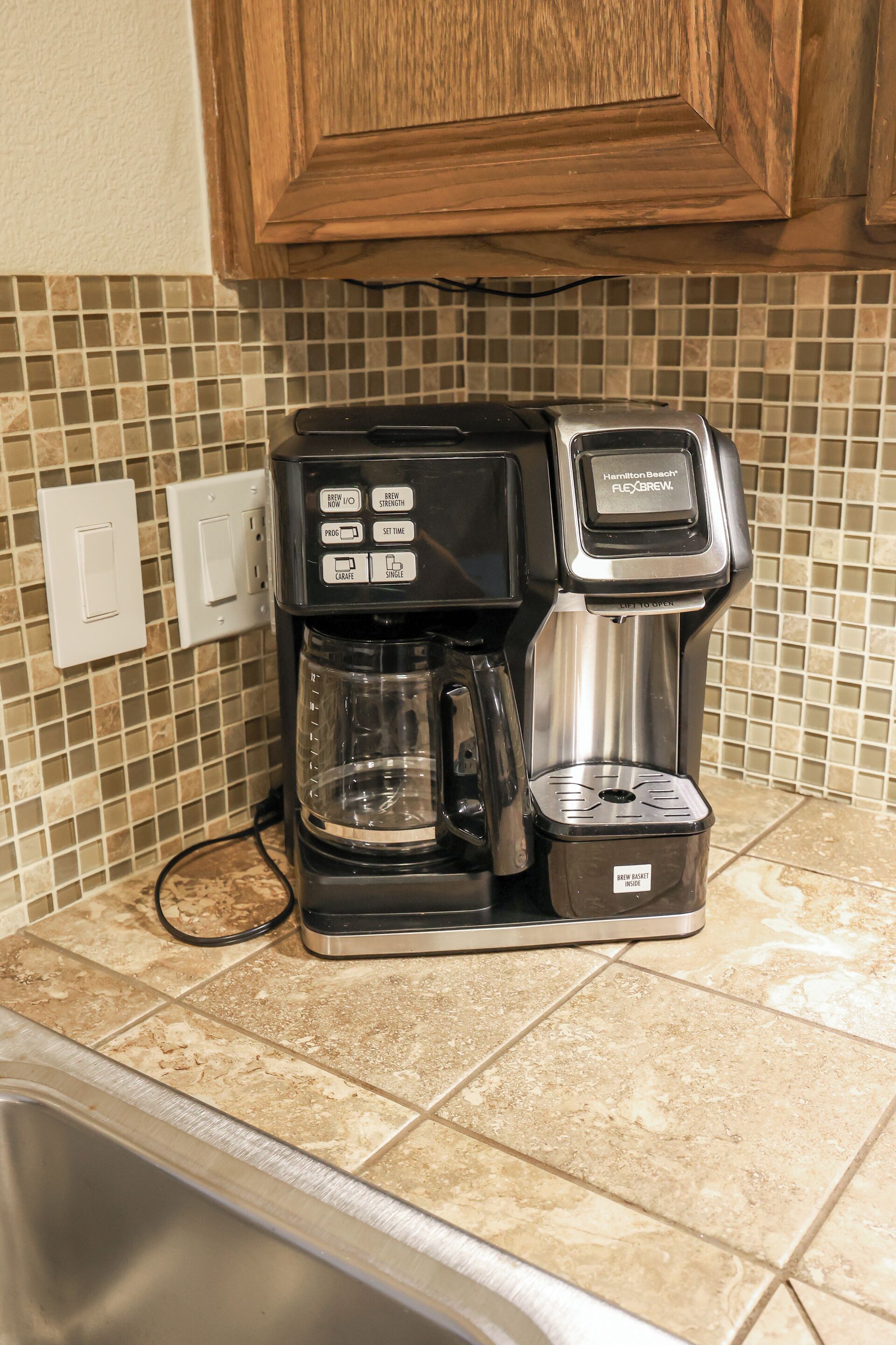 A Hamilton Beach Flexbrew coffee maker on a kitchen counter with a tiled backsplash and wooden cabinets.