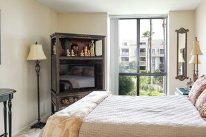 A bedroom with a large bed, a dresser with decorative items, a standing lamp, and a window with a view of buildings and palm trees. - Bahama Hideaway (Oceanside)