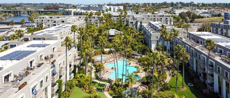 Un hotel de varios pisos, con una piscina central rodeada de palmeras y paneles solares en los tejados.