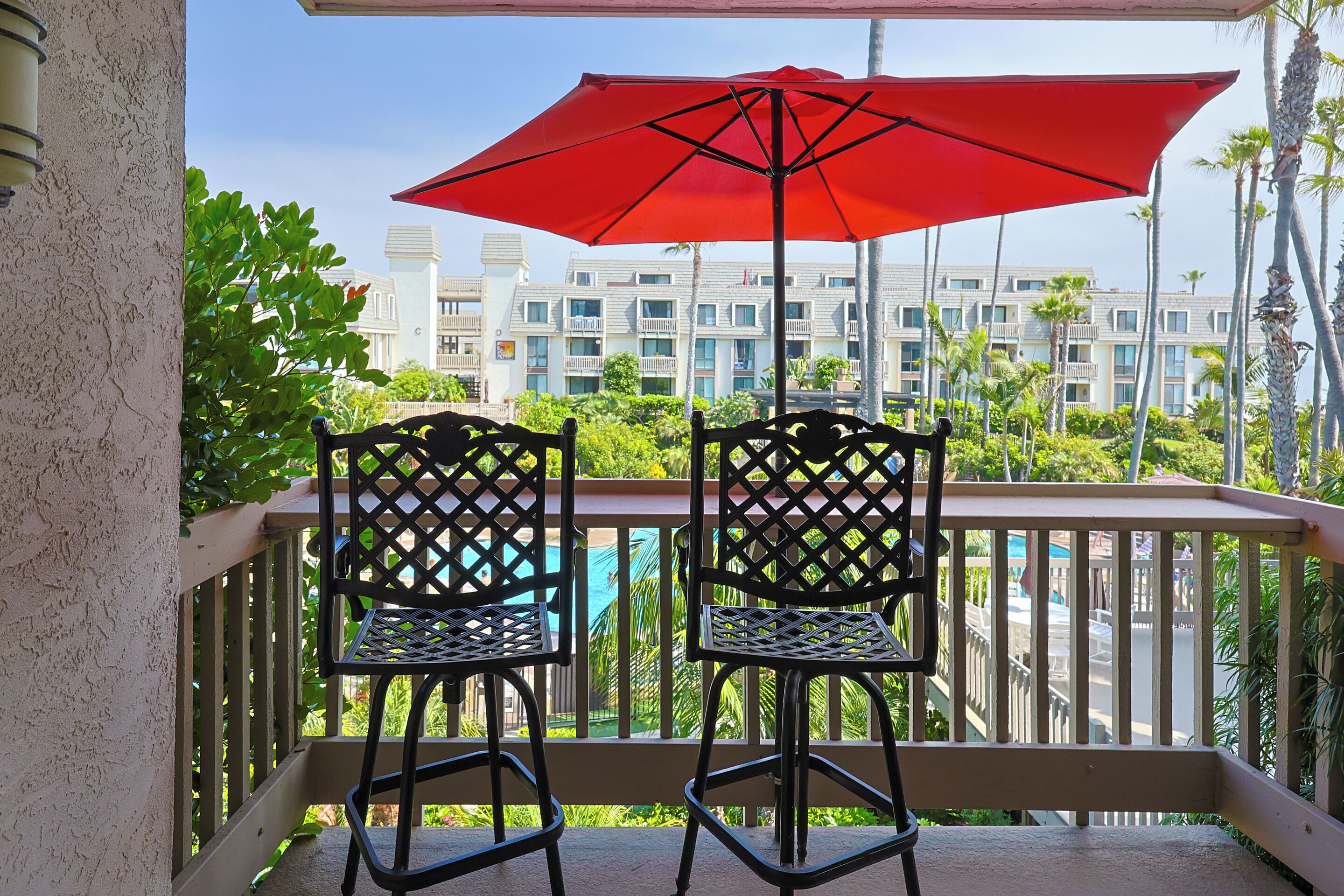 Two black chairs with a red umbrella on a balcony overlooking a pool and a building with balconies.
