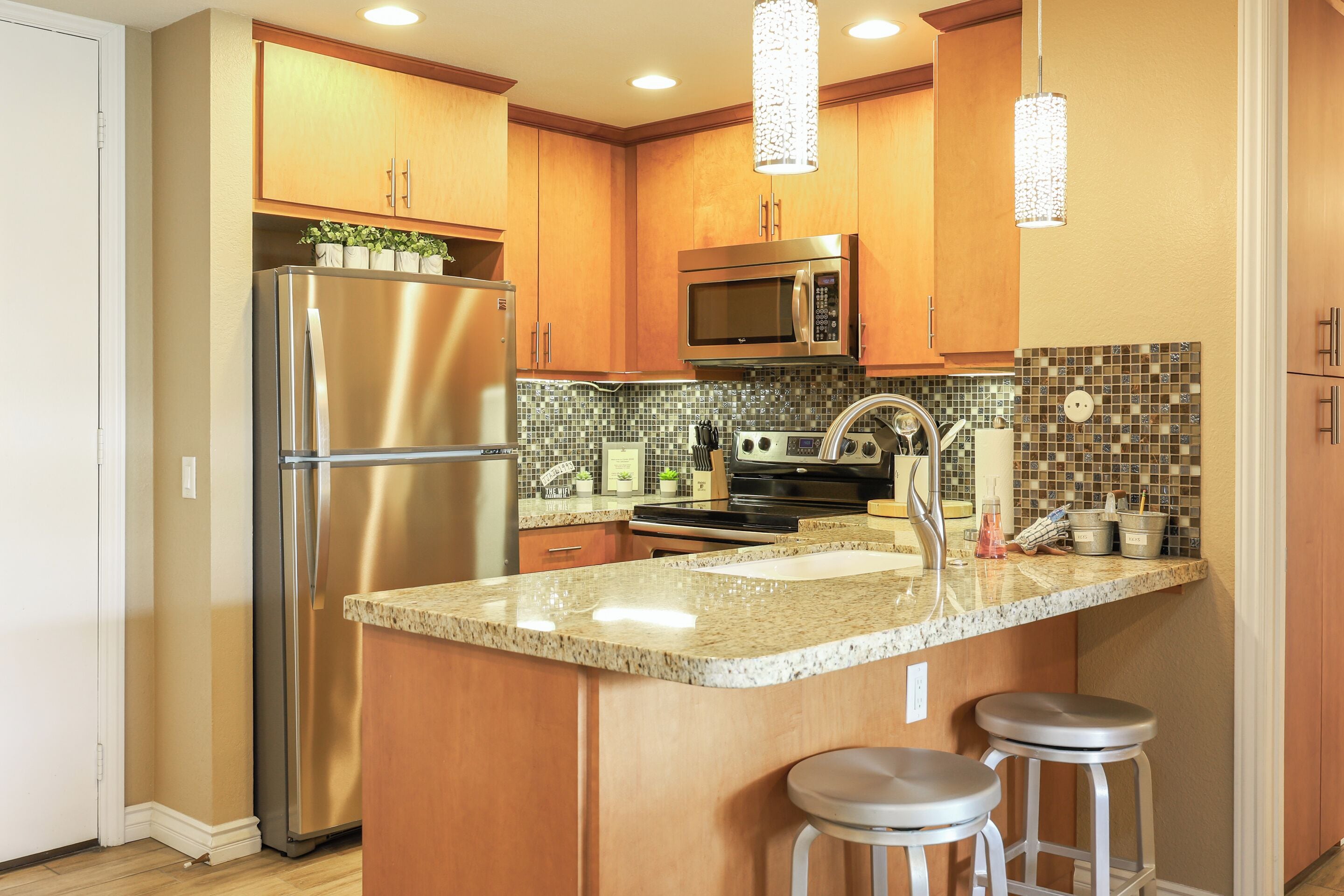 A modern kitchen with wooden cabinets, stainless steel appliances, a granite countertop, and two bar stools.