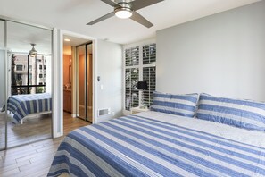 A bedroom with a large bed, a ceiling fan, a sliding glass door leading to a balcony, and a view of a building across the street. - Saphire Sands (Oceanside)
