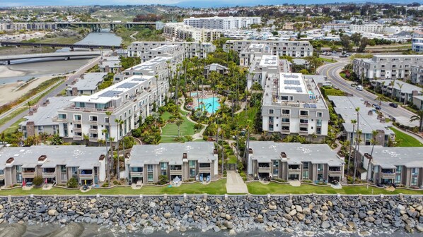 A modern residential complex with multiple apartment buildings, a central swimming pool, and a bridge in the background. - Tropical Beach Retreat (Oceanside)