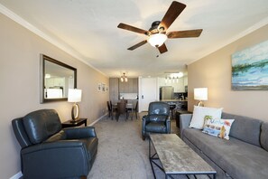 A living room with a ceiling fan, a gray sofa, a black leather chair, a coffee table, and a dining area with a table and chairs. - Harbor Horizon Retreat (Oceanside)