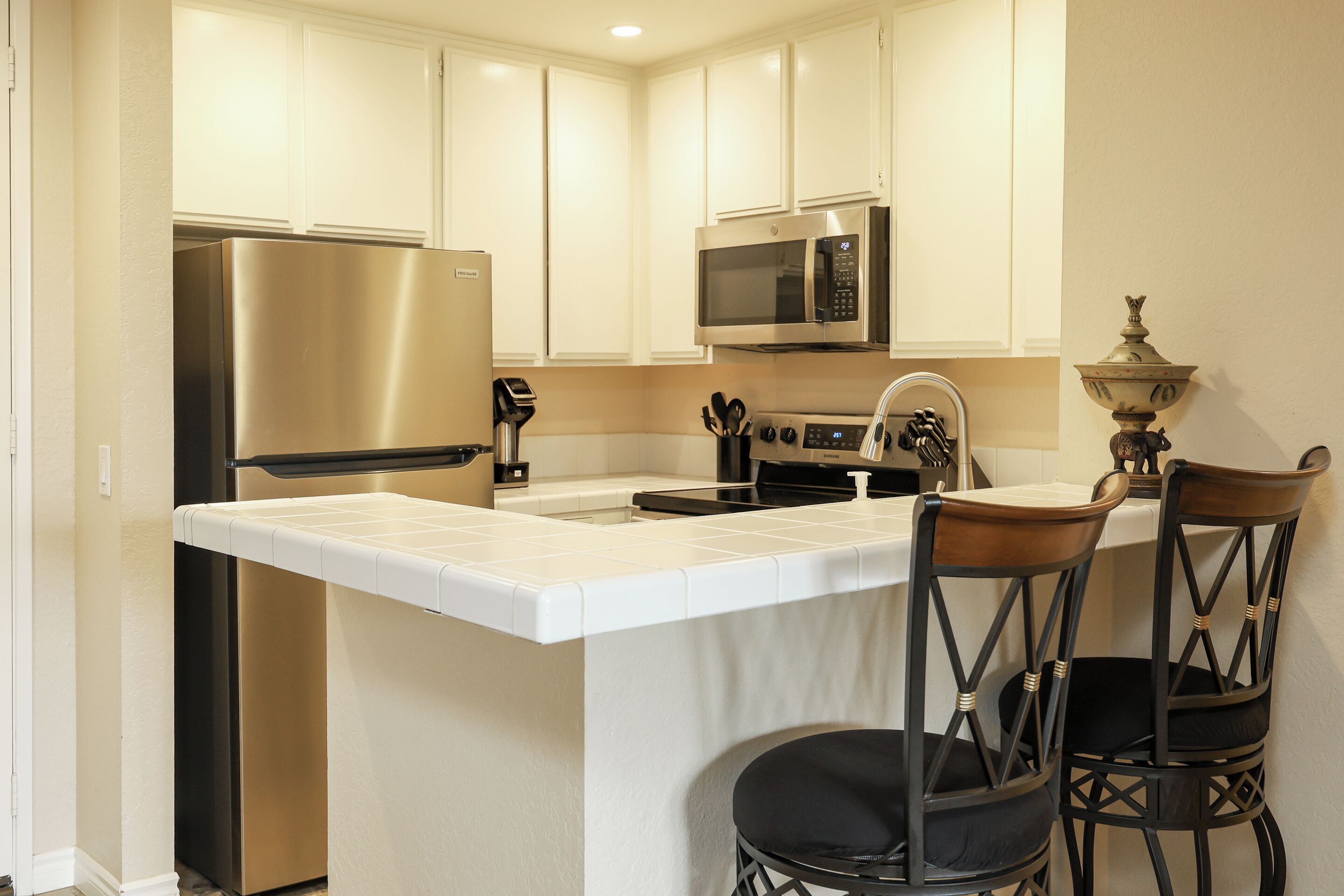 A kitchen with white cabinets, stainless steel appliances, a white countertop, and two black upholstered chairs.