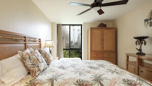 A bedroom with a bed, a ceiling fan, a window with a view of palm trees, and a wooden cabinet. - Enjoy Beautiful Views in This New One Bedroom Unit (Oceanside)