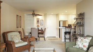 A living room with a green sofa, wicker armchair, and a coffee table. There is a dining area with a table and chairs, and a kitchen area with a refrigerator and microwave. - Enjoy Beautiful Views in This New One Bedroom Unit (Oceanside)