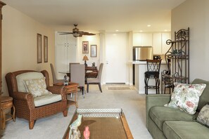 A living room with a green sofa, wicker armchair, and a coffee table. There is a dining area with a table and chairs, and a kitchen area with a refrigerator and microwave. - Enjoy Beautiful Views in This New One Bedroom Unit (Oceanside)