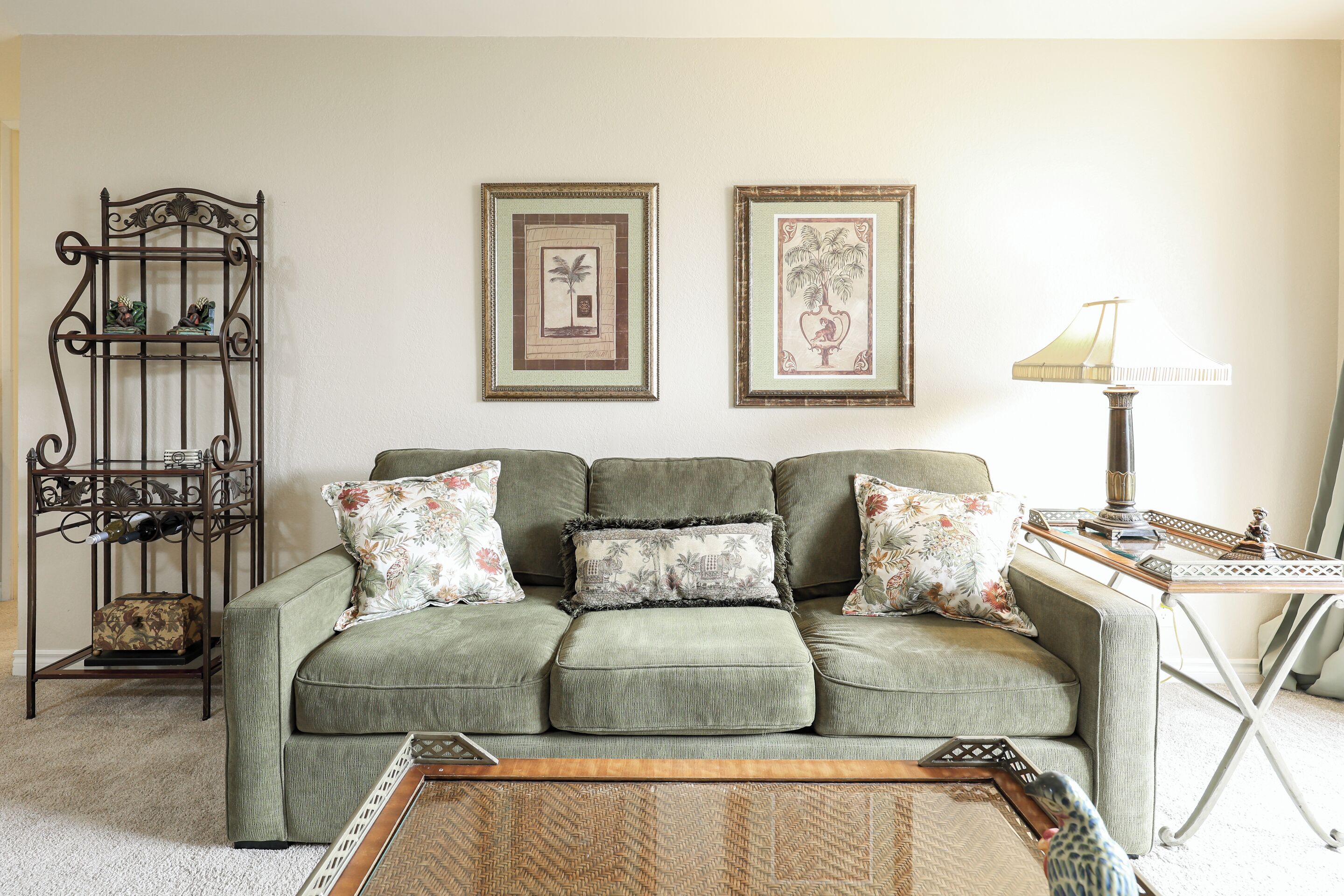 A green sofa with floral cushions, a metal rack with bottles, and two framed pictures on the wall.