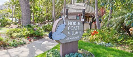 Un panneau indiquant l’entrée de North Coast Village, situé au 999 North Pacific, entouré d’une végétation luxuriante et d’un jardin bien entretenu.