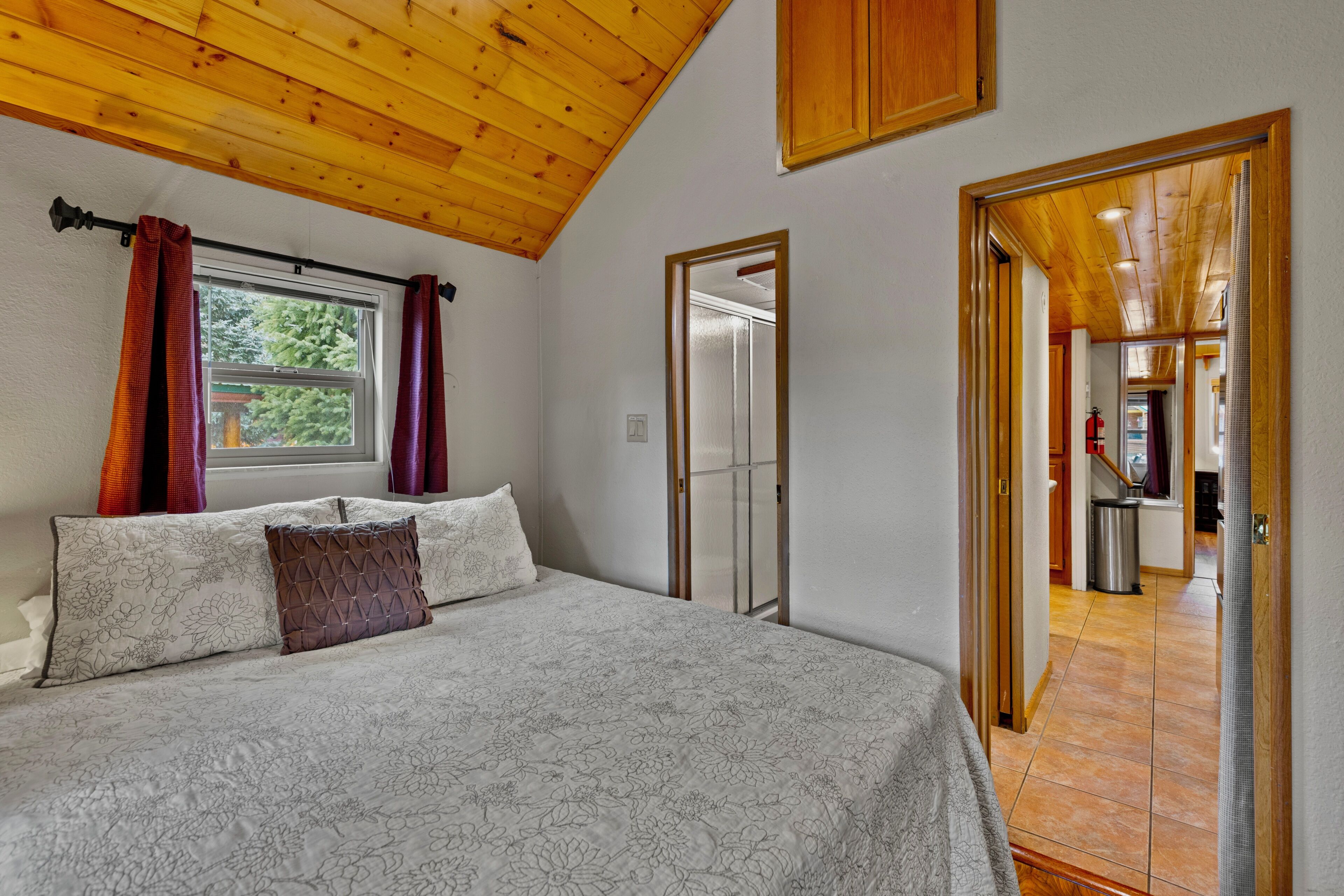 A bedroom with a wooden ceiling, a bed with floral bedding, a window with curtains, and a door leading to another room.