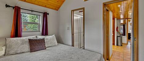 A bedroom with a wooden ceiling, a bed with floral bedding, a window with curtains, and a door leading to another room.