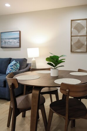 A dining area with a round wooden table, four chairs, a potted plant, a lamp, and a framed picture on the wall. - Stylish 1-bedroom Ocean Unit: Renovated With Peaceful Garden Views (Oceanside)