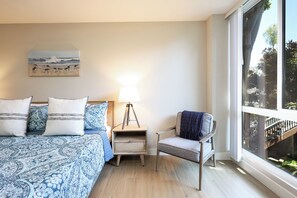 A bedroom with a bed, bedside table, lamp, chair, and a painting on the wall. - Horizon Sweet (Oceanside)