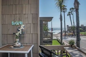 A wooden wall with a 'Surfs Up' sign, a table with a vase of flowers, and a view of the beach with palm trees. - The Blue Pearl (Oceanside)