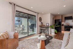 A modern living room with a large sliding glass door leading to a balcony, a dining area with a bar, and a kitchen with a black refrigerator. - Romantic Lakefront Condo F5 + Pool - Wapato Chelan (Manson)