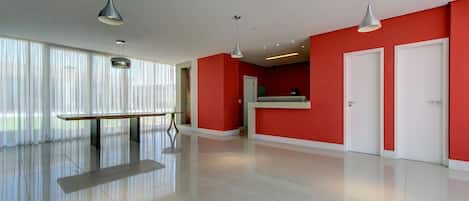 A spacious, modern hotel lobby with a red accent wall, a reception desk, and a large window with sheer curtains.