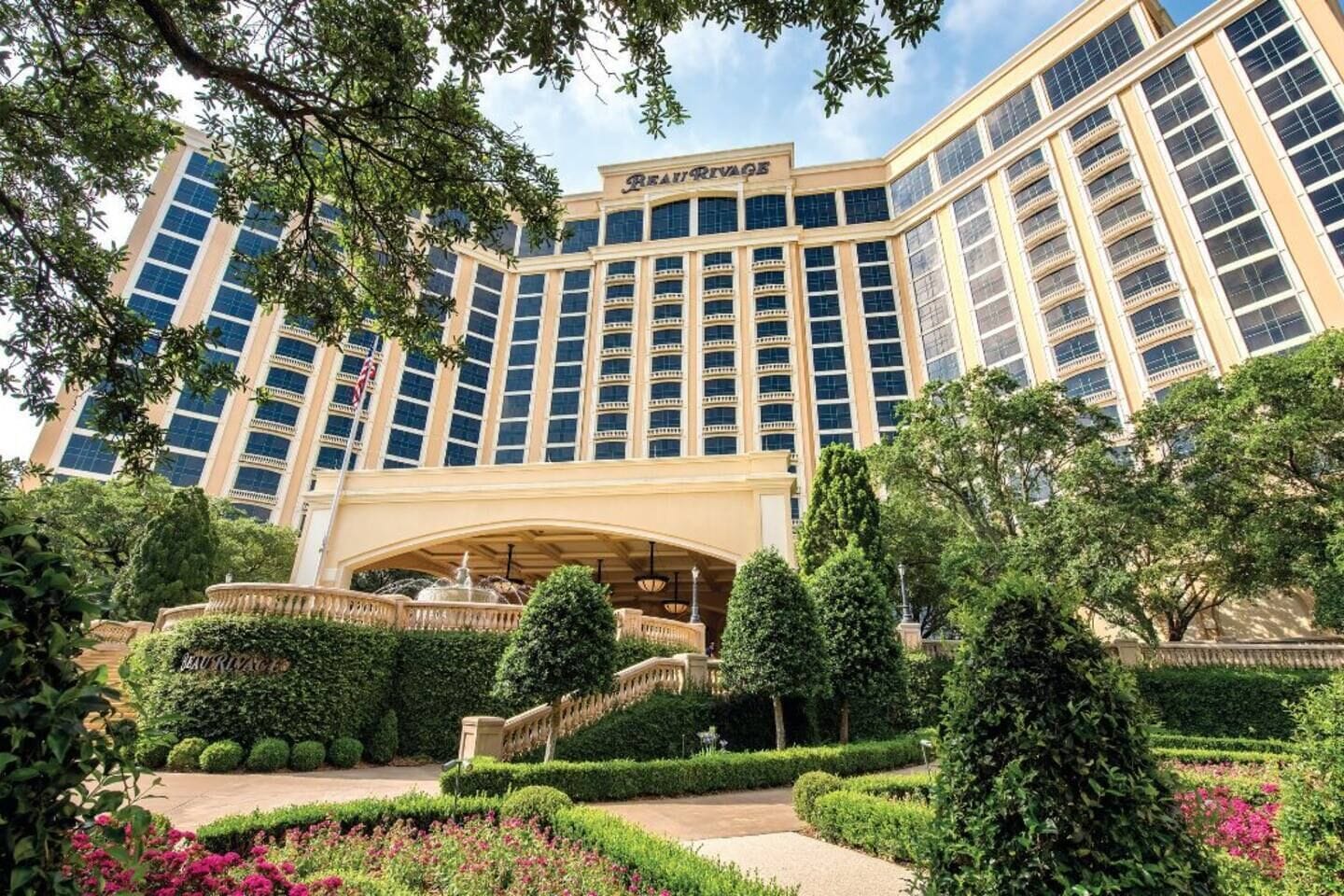 A large hotel with a central entrance, multiple floors, and a well-maintained garden with hedges and flowers.