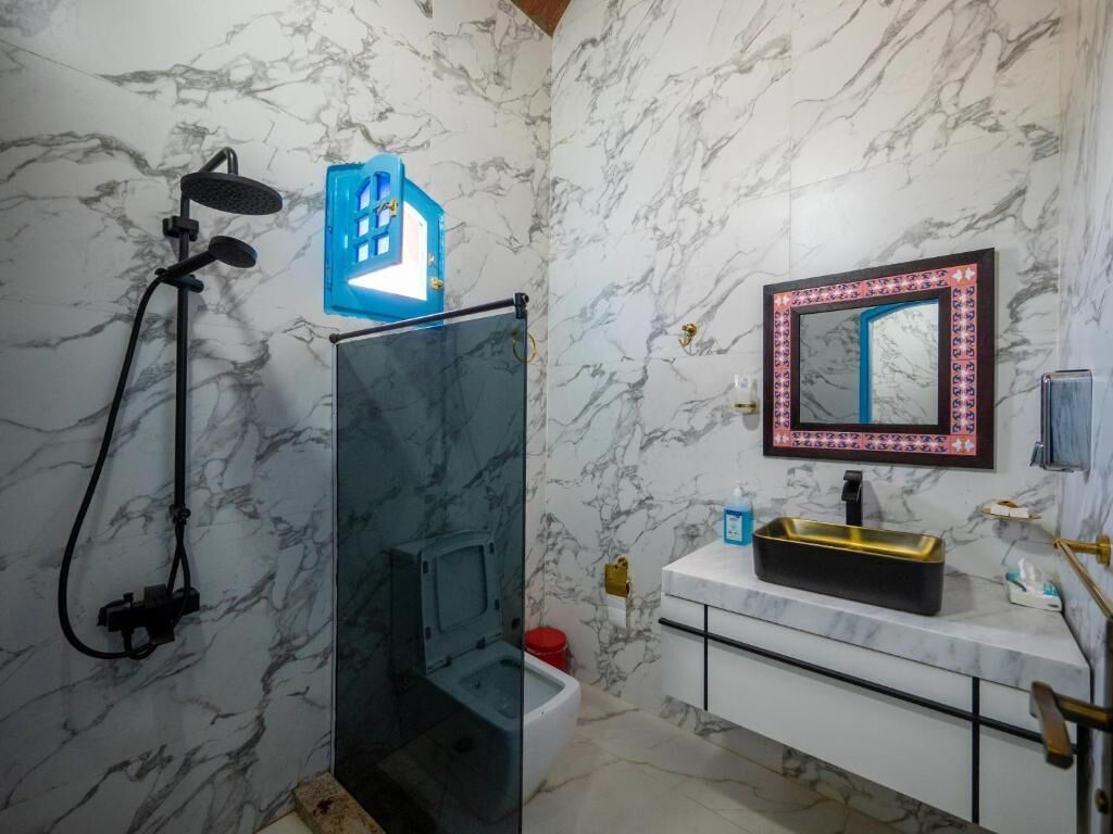 Une salle de bain avec un mur en marbre, une douche avec une porte en verre, un lavabo avec un receveur doré et un miroir avec un cadre décoratif.