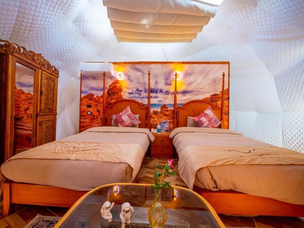 A room with two beds, a wooden cabinet, a glass table with a vase, and a sunset mural on the wall.