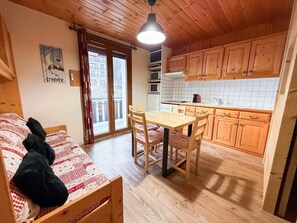 A cozy room with a bed, a dining table with chairs, a kitchen with wooden cabinets, and a large window.
