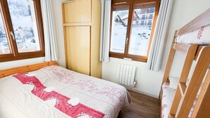 A bedroom with a bed, wooden wardrobe, two windows with curtains, a radiator, and a view of a snowy landscape. - Les Balcons de St So Apartment 1 – Mountain View, 2 Bedrooms, Near Slopes (Saint-Sorlin-d'Arves)